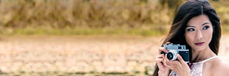 Vintage style panorama web banner header of beautiful Asian Chinese girl or young woman wearing summer dress outside in green environment taking pictures or photographs in Autumn or Fall with a retro cameraの写真素材