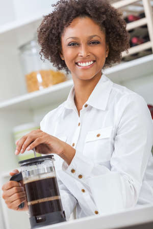 Beautiful happy mixed race African American girl or young woman with perfect teeth in her kitchen, making coffee with a French press or cafetiereの写真素材