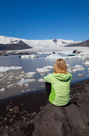 Climate Change Global Warming Concept of young woman female hiker wearing a green jacket sitting on a rock looking out on a melting glacier and icebergs in Icelandの写真素材
