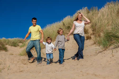 Happy family of mother, father and two sons, man woman children walking holding hands, having fun in the sand dunes of a sunny beachの写真素材