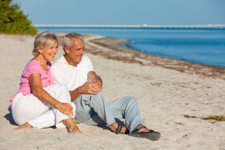 Happy senior man and woman couple sitting, smiling and laughing on a sunny beachの写真素材