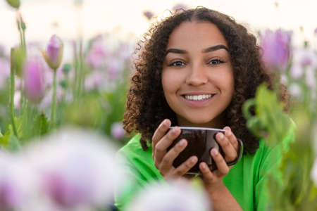 Beautiful happy mixed race biracial African American girl teenager female young woman smiling drinking coffee or tea outdoors in a field of pink and purple poppies flowersの写真素材