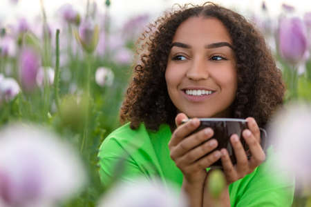 Mixed race beautiful African American biracial girl teenager female young woman drinking cup of tea or coffee wearing a green t-shirt in field of pink and purple flowers smiling with perfect white teethの写真素材
