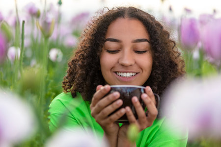 Mixed race beautiful African American biracial girl teenager female young woman drinking cup of tea or coffee wearing a green t-shirt in field of pink and purple flowers smiling with perfect white teethの写真素材