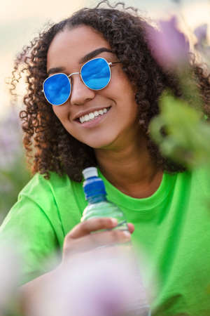 Mixed race beautiful African American biracial girl teenager female young woman wearing blue sunglasses and drinking a bottle of water in field of pink flowers poppies at sunset or sunrise smiling with perfect white teethの写真素材