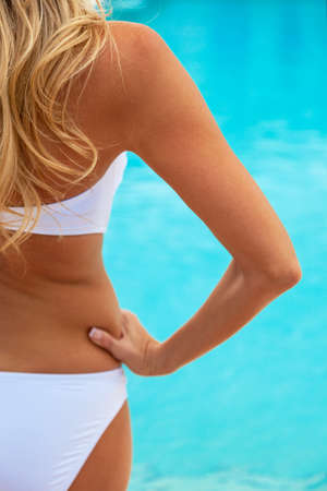 Rear view of beautiful sexy young blonde woman wearing a white bikini standing by a turquoise blue swimming pool. Spa, vacation, healthy living and health club concept.の写真素材