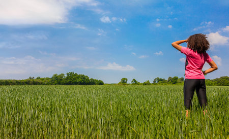 Rear view of young woman girl female runner jogger running standing in green field with blue skyの写真素材