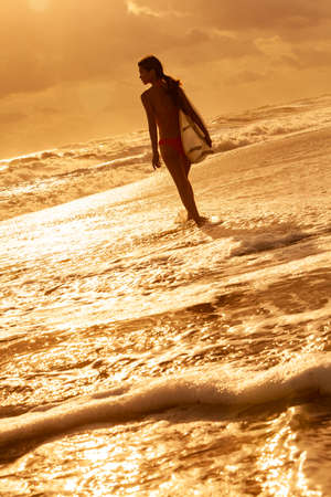 Rear view of beautiful sexy young woman surfer girl in bikini with white surfboard on a beach at sunset or sunriseの写真素材