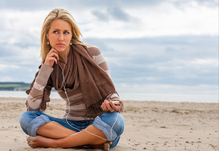 Beautiful thoughtful sads depressed young blond woman listening to music on a beach with her smartphone cell phone and headphonesの写真素材