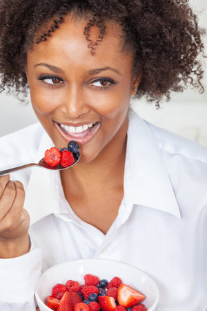 African American biracial girl female young woman with perfect teeth eating a bowl of fresh fruit, strawberries, raspberries and blueberries. Healthy eating and lifestyle concept.の写真素材