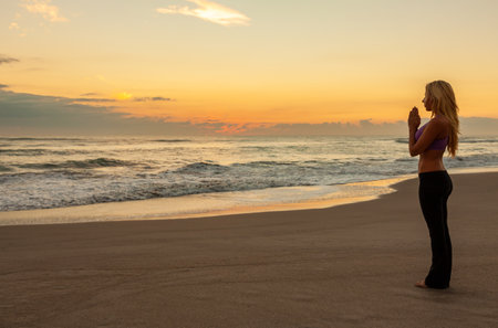 Young fit blonde woman alone practicing yoga on a beach at sunrise or sunsetの写真素材