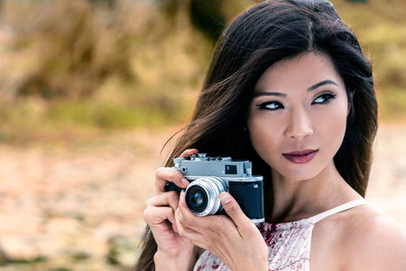 Vintage style portrait of beautiful Asian Chinese girl or young woman wearing summer dress outside in green environment taking pictures or photographs in Autumn or Fall with a retro cameraの写真素材