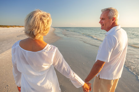 Happy senior man and woman old retired couple walking and holding hands on a beach at sunsetの写真素材