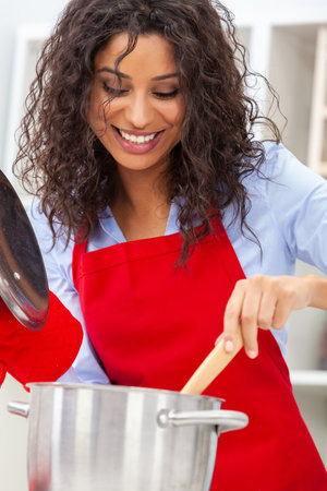 Beautiful Latina girl or young woman looking happy wearing red apron, cooking holding metal pot in her kitchen at homeの写真素材