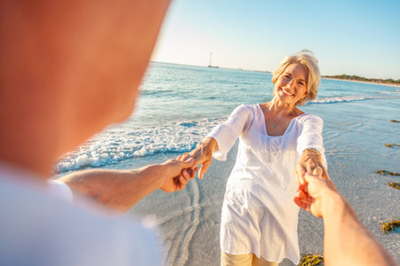 Happy senior man and woman old retired couple dancing and holding hands on a beach with bright clear blue skyの写真素材