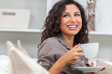 Beautiful young Latina Hispanic woman smiling, relaxing and drinking a cup of coffee or tea at homeの写真素材