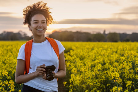 Beautiful mixed race African American girl teenager female young woman photographer smiling and happy taking photographs with a camera in a field of yellow flowers at sunset or sunriseの写真素材