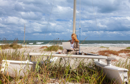 Beautiful successful young woman sitting barefoot on the deck of a small catamaran sailing boat using her laptop computer remote working on line from homeの写真素材