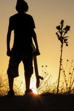 young skater holding his skateboard in his hand against the light watching a beautiful sunset next to a plant with spikesの写真素材