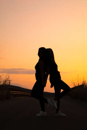 two young brunette girl posing a road in the middle of the field at sunset, silhouetteの写真素材