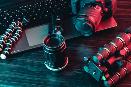 A workspace with laptop, modern camera, lens, tripod and a black pen on a wood background. Freelance or creative man or woman. Red and blue lightの写真素材
