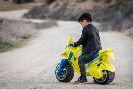 A happy little boy driving a toy motorcycle, dressed in a leather biker jacket on a country roadの写真素材
