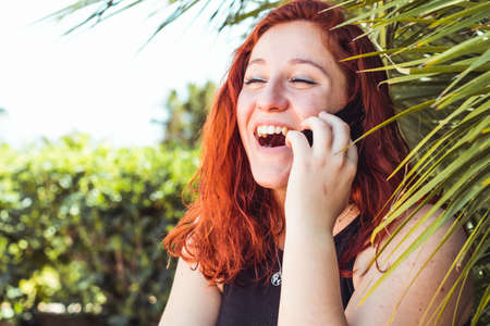 Close up of smiling attractive girl with redhead talking on mobile phone while standing outdoorsの写真素材