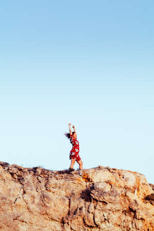 Young girl with red dress on cliff's edge with raised hands and looking to a skyの写真素材