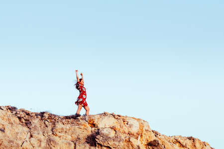Young girl with red dress on cliff's edge with raised hands and looking to a skyの写真素材