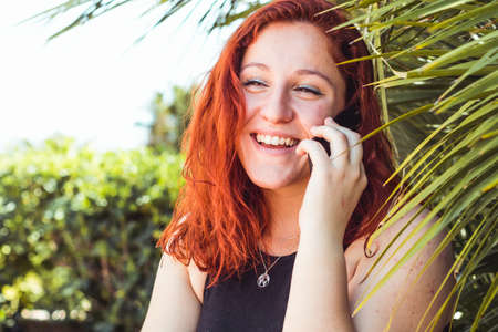 Close up of smiling attractive girl with redhead talking on mobile phone while standing outdoorsの写真素材