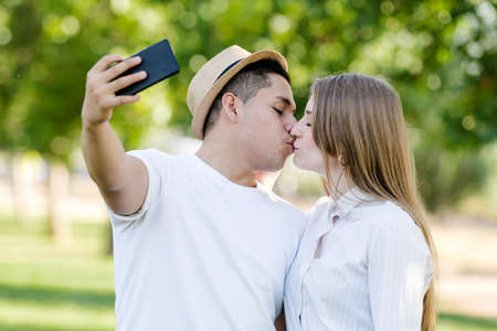 Young couple kissing and taking a selfie on their mobile in the park. Latin man and Caucasian womanの写真素材