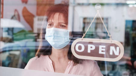 Woman with face mask changing closed to open sign on window looking outsideの写真素材