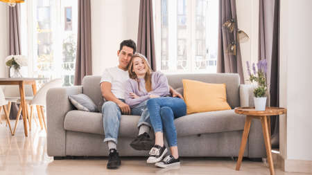 Portrait of young couple smiling and looking at the camera while sitting on the couch at home.の写真素材