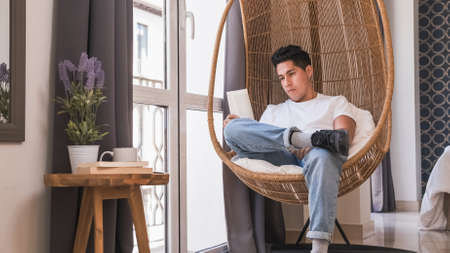 Portrait of young man reading a book while sitting in a comfortable chair at home.の写真素材
