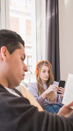 Young couple relaxing and sitting on couch at home while reading a book and listening music with headphonesの写真素材