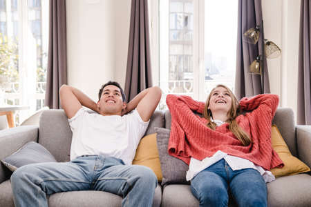 Portrait of young couple putting hands behind head and relaxing while sitting on sofa at home.の写真素材