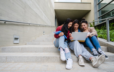 Group of Asian female students using a laptop and studying together while sitting on the stairs of...