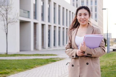 Asian university student looking at the camera and holding folders while standing outdoors at the...