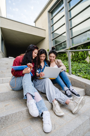Group of Asian female students using a laptop and studying together while sitting on the stairs of the university campus.の写真素材