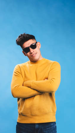 Man in sunglasses smile while posing with his arms crossed over an isolated blue background.の写真素材