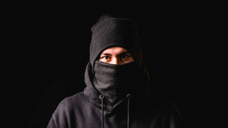 Thief in black balaclava and black cap looking at camera while standing against dark background.の写真素材