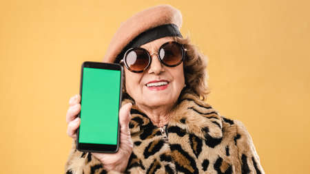 Stylish senior woman showing a mobile phone with green while standing over an isolated background.の写真素材