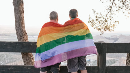 Back view of a male couple embracing each other while holding an LGBT rainbow flag outdoors.