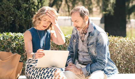 Senior couple looking sad and disappointed while using a laptop in a sunny parkの写真素材