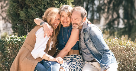 Two senior women and one senior man are sitting in a sunny park, smiling and hugging each otherの写真素材