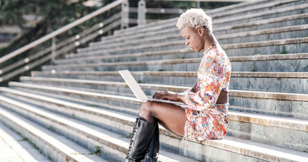 Transgender woman sitting on exterior steps using a laptop, remote workerの写真素材