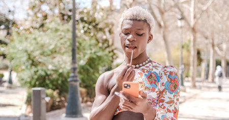 Transgender woman applying herself lipstick looking at mobile phone while standing outdoors.の写真素材