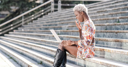 Transgender woman sitting on exterior steps using a laptop, remote workerの写真素材