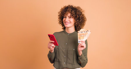 Excited woman with cash and looking phone in beige studioの写真素材