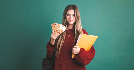 Smiling female university student holding books and fanned out 50 euro banknotesの写真素材